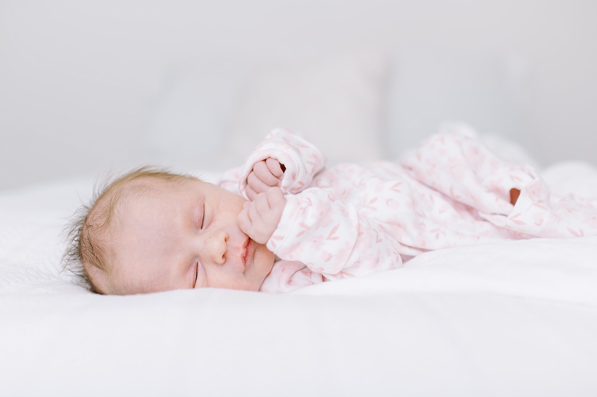 Newborn baby girl sleeps on her side wearing pink pajamas while laying on white bedding.