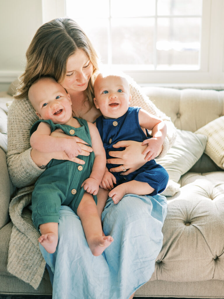 Mother holds twin babies on lap as sun shines in behind them by Emilie Phillipson Photography. 