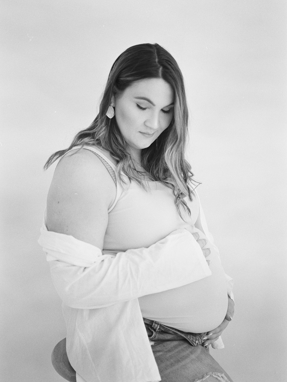 Black and white film photograph of mother looking down and holding her belly in her hands with her white sweater draped down her shoulder with jeans unbottoned at the top