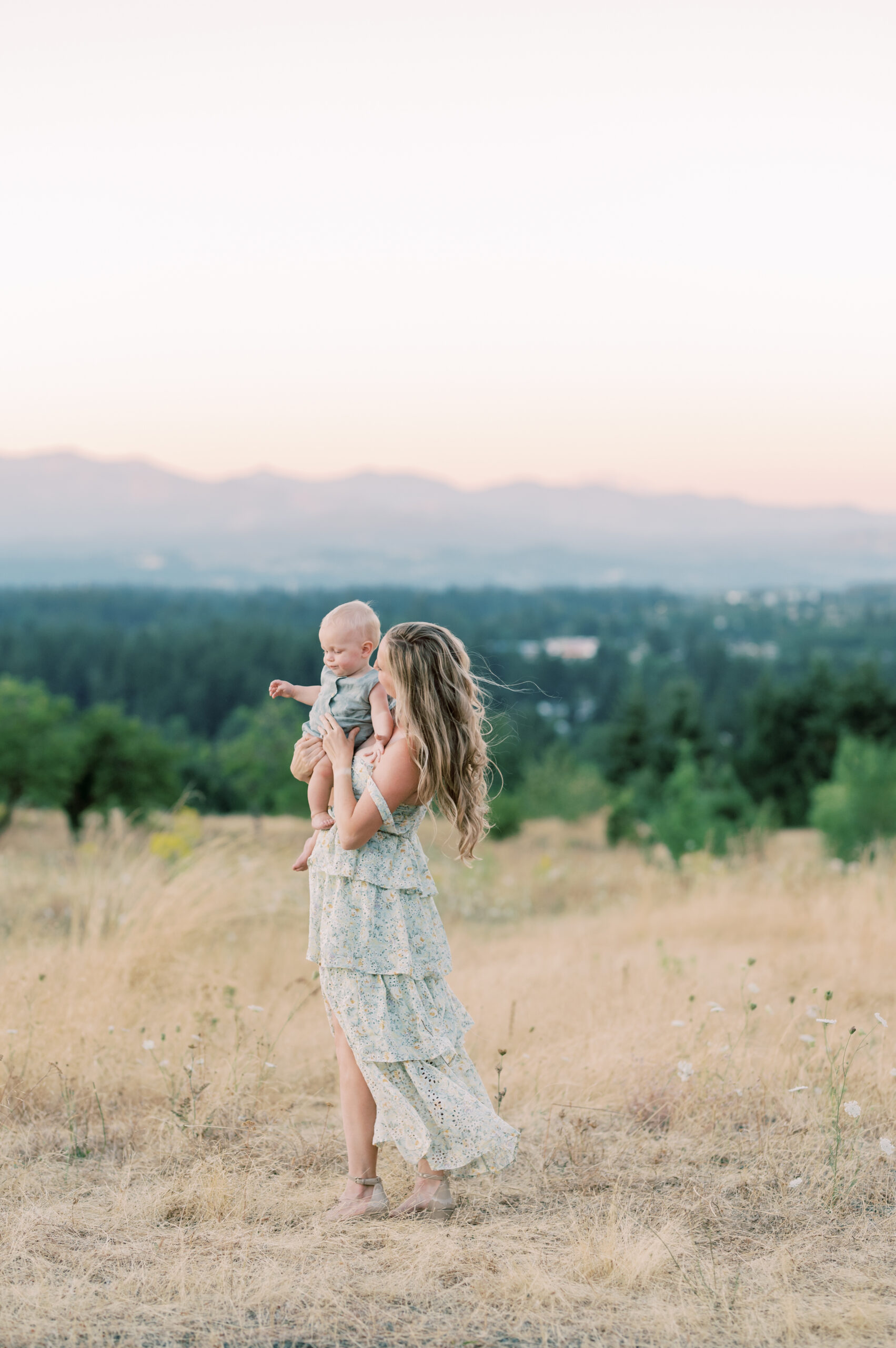 Mother with long blonde hair soft curls wearing a long flowy tiered mint green dress holding her young child, kissing his hand, standing in an empty field overlooking the city of portland with green trees, sparkling lights, and a colorful sunset of blues, oranges, and pink skylines.