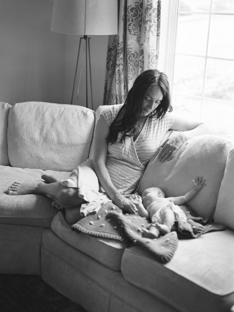 Black and white film image of Mother with dark long hair and light green dress sits on yellow sofa and holds hand of newborn baby who is laying next to her on sofa by Portland Newborn Photographer Emilie Phillipson Photography. 
