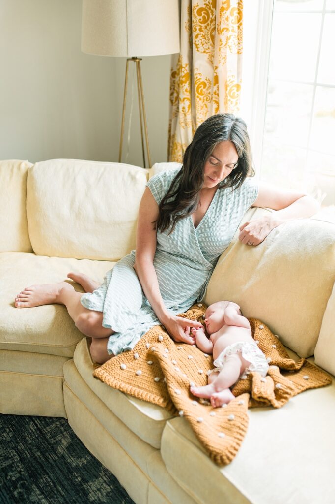Mother with dark long hair and light green dress sits on yellow sofa and holds hand of newborn baby who is laying next to her on sofa by Portland Newborn Photographer Emilie Phillipson Photography. 
