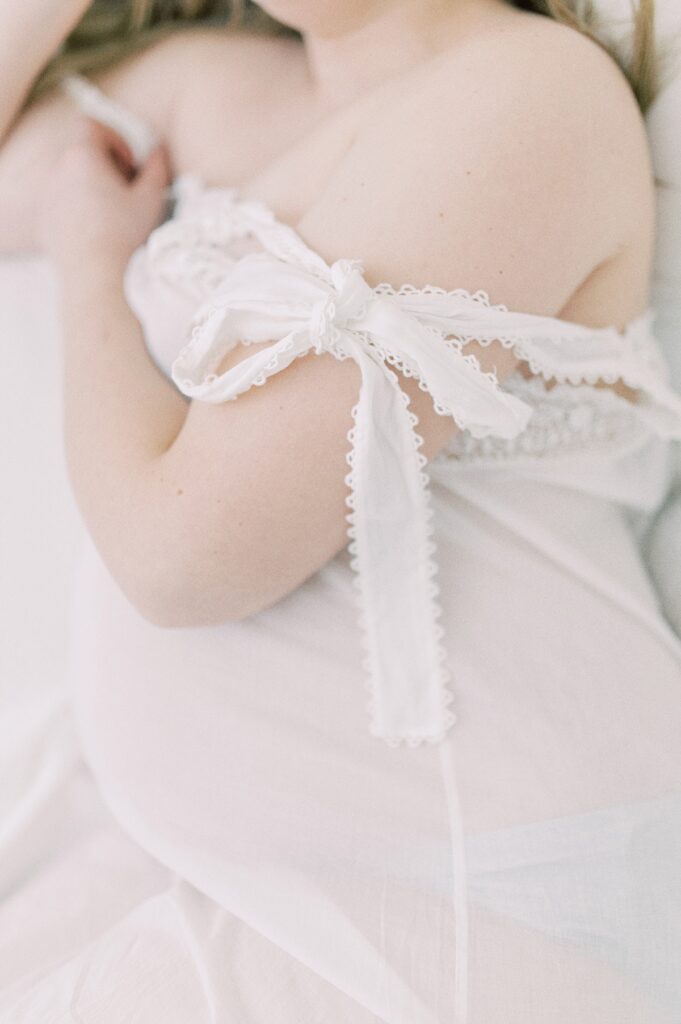 Pregnant woman wearing white cotton gown with should strap partially untied and draped down her arm laying down by Portland Maternity Photographer Emilie Phillipson Photography. 