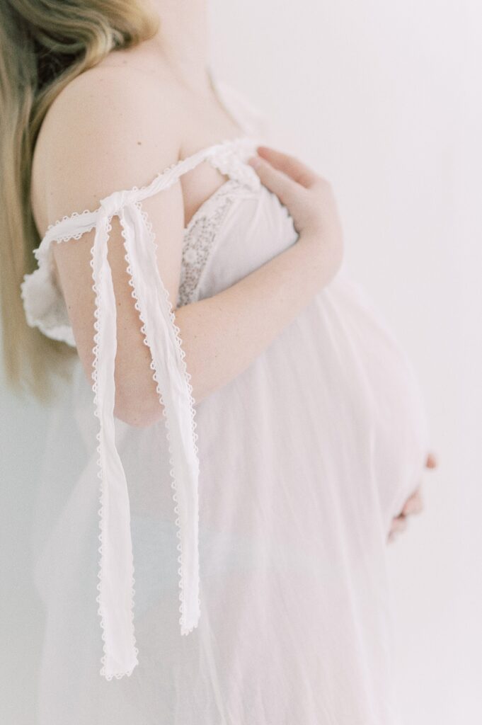 Pregnant woman wearing white cotton gown with should strap partially untied and draped down her arm with hands holding up gown and on belly by Portland Maternity Photographer Emilie Phillipson Photography. 