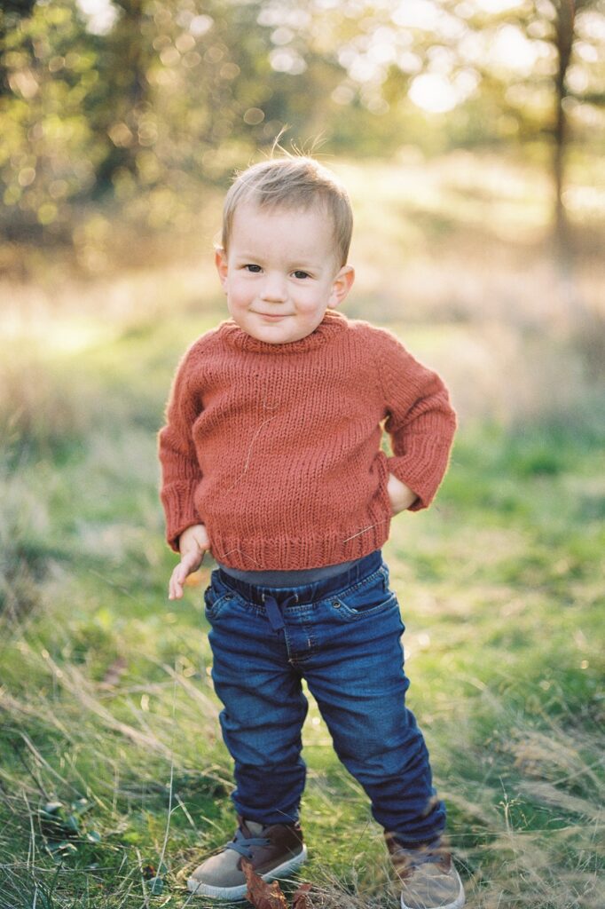 Portrait image of toddler boy with dark brown eyes wear rust orange sweater and blue jeans standing in grassy field with light sparkling through background during Fall Family Session in Portland Oregon with Family Photographer Emilie Phillipson Photography.