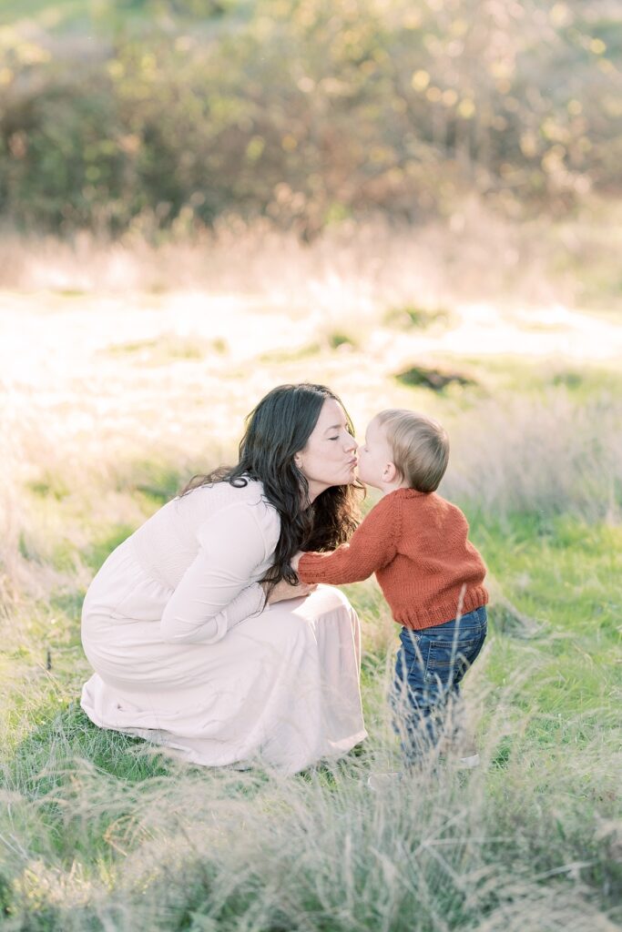 Mother wearing long beige dress kneels down to kiss toddler son on lips wearing jeans and rust orange sweater in grassy light field during Fall Family Session in Portland Oregon by Emilie Phillipson Photography. 