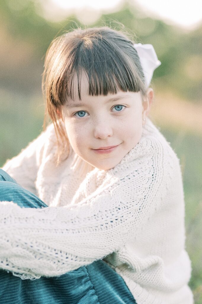 Portrait image of young girl with blue eyes looking directly at camera view wearing beige sweater and teal velvet skirt, with sunlight shining on her brown hair and through background during Fall Family Session in Portland Oregon with Family Photographer Emilie Phillipson Photography.