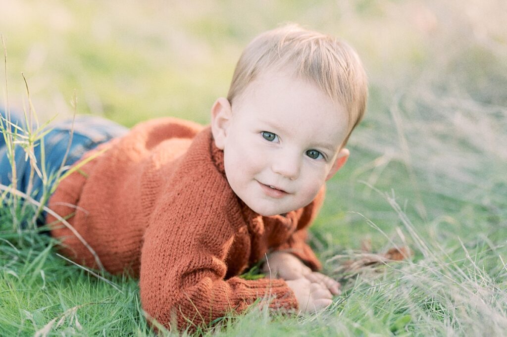 Portrait image of toddler boy with dark brown eyes wear rust orange sweater and blue jeans laying in grassy field with light sparkling through background during Fall Family Session in Portland Oregon with Family Photographer Emilie Phillipson Photography.