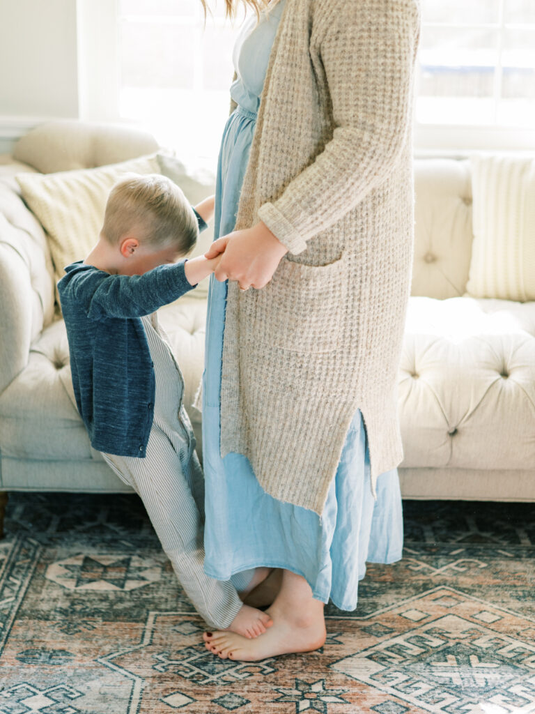 Young boy stands on mother's feet wearing sweaters as sun shines in behind them from by Portland Family Photographer Emilie Phillipson Photography. 