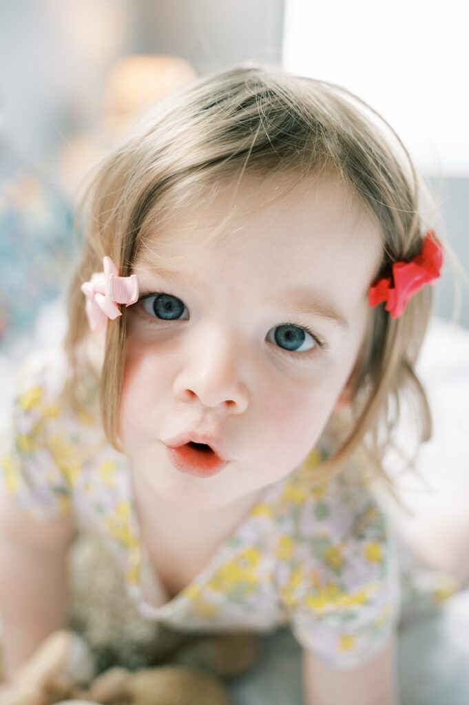 Close up of toddler sister with silly face and hair bows in hair that are twisted with big blue eyes looking right at the camera by  Emilie Phillipson Photography